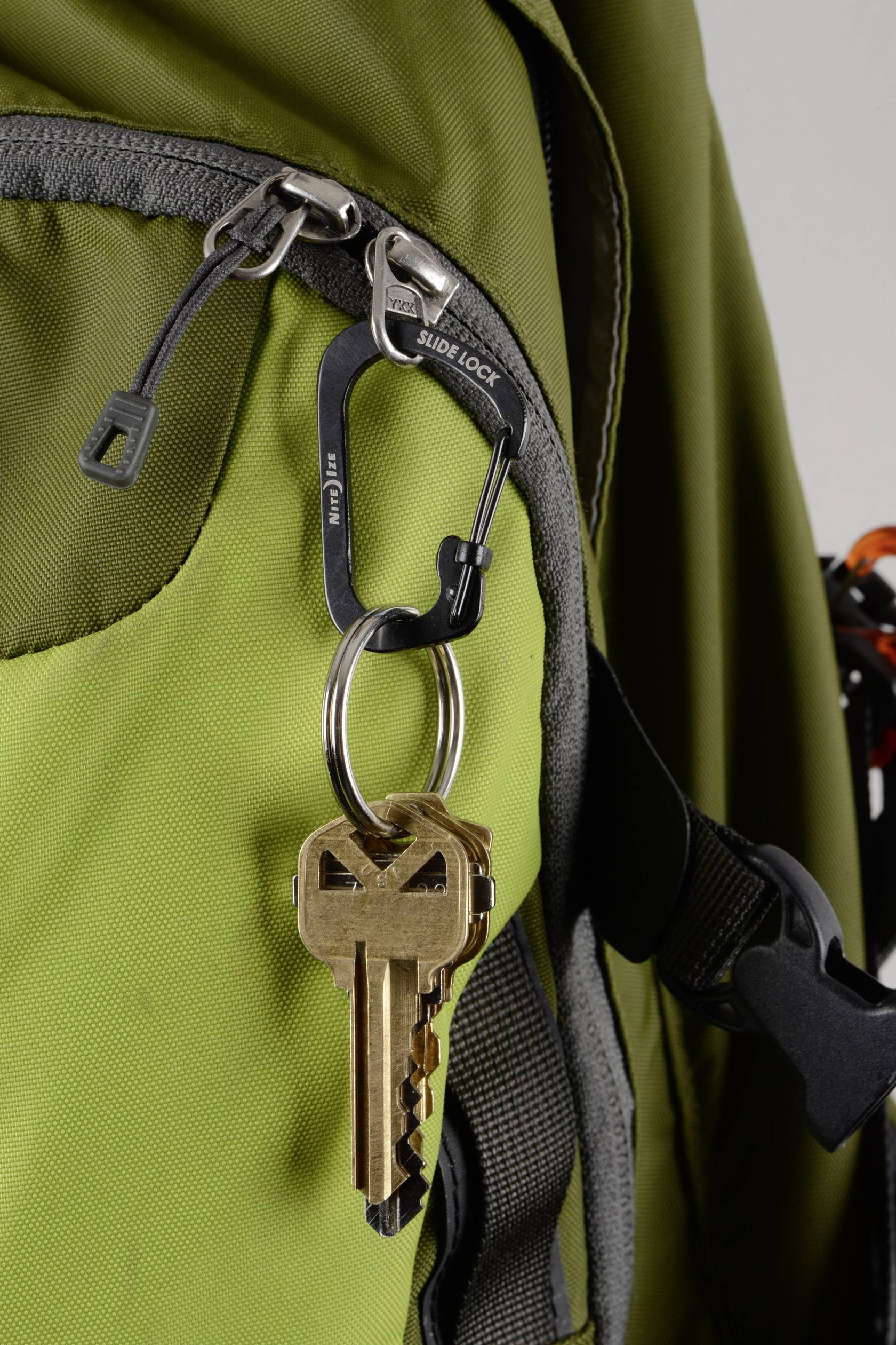 Ein goldener Schlüssel hängt an einem silbernen Karabinerhaken, der an einem Reißverschluss eines grünen Rucksacks befestigt ist.