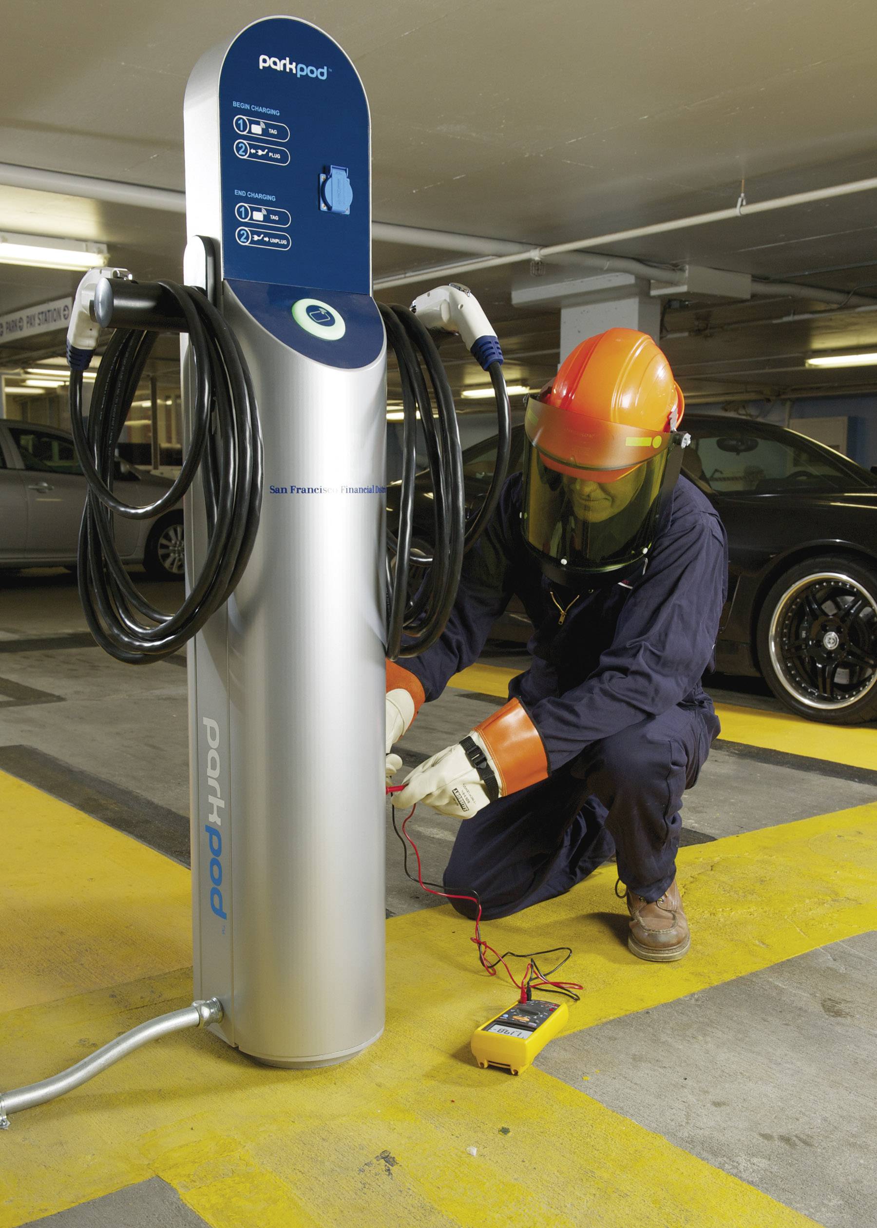 Ein Techniker mit Schutzkleidung überprüft eine Elektroauto-Ladestation in einer Tiefgarage. Ein Messgerät liegt auf dem Boden.