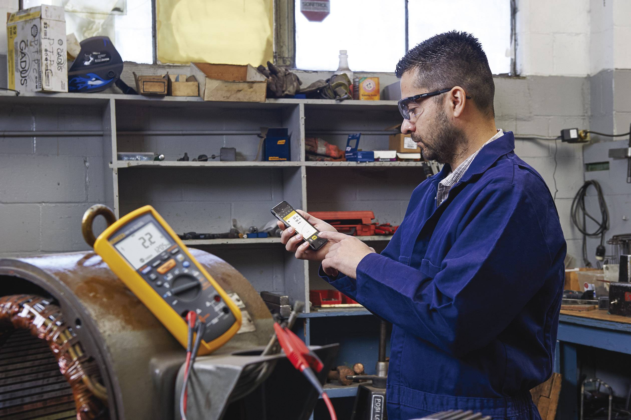 Ein Mann in blauer Arbeitskleidung misst mit einem Multimeter die Elektronik. Im Hintergrund sind Werkzeuge und Regale sichtbar.