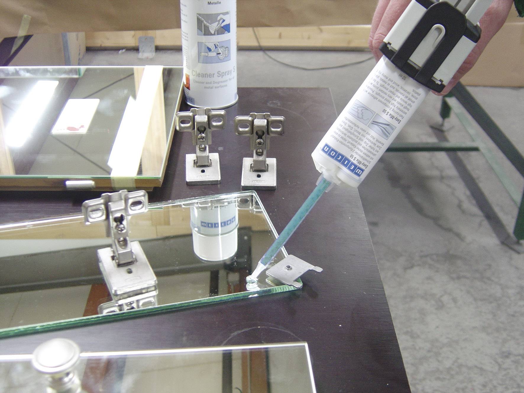 Eine Person trägt mit einer Kartuschenpistole Klebstoff auf den Rand eines Spiegels auf. Im Hintergrund sind weitere Spiegelteile und Scharniere zu sehen.