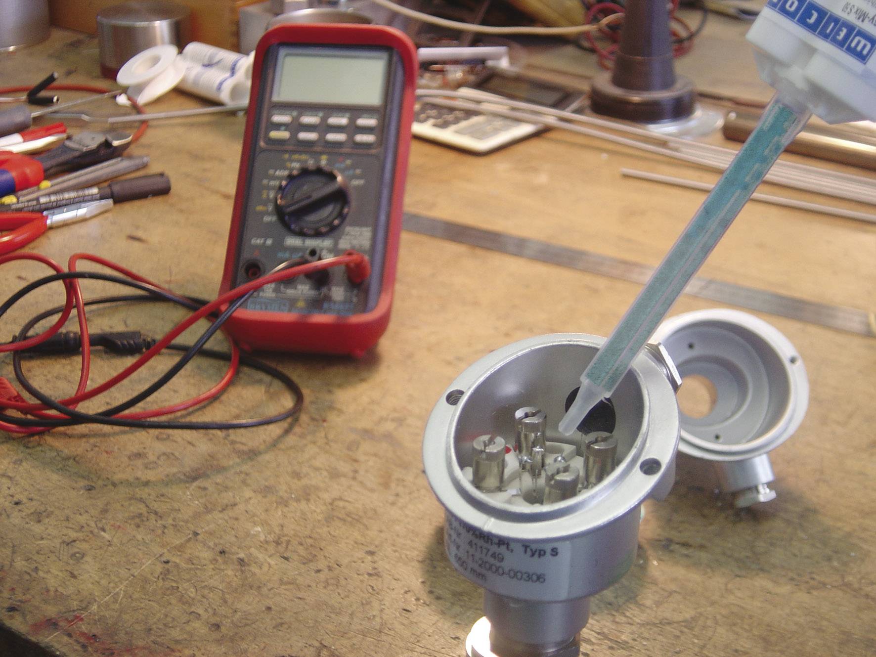 Eine Hand füllt eine elektrische Komponente mit Silikon auf einer Werkbank, neben einem eingeschalteten Multimeter und Werkzeugen.