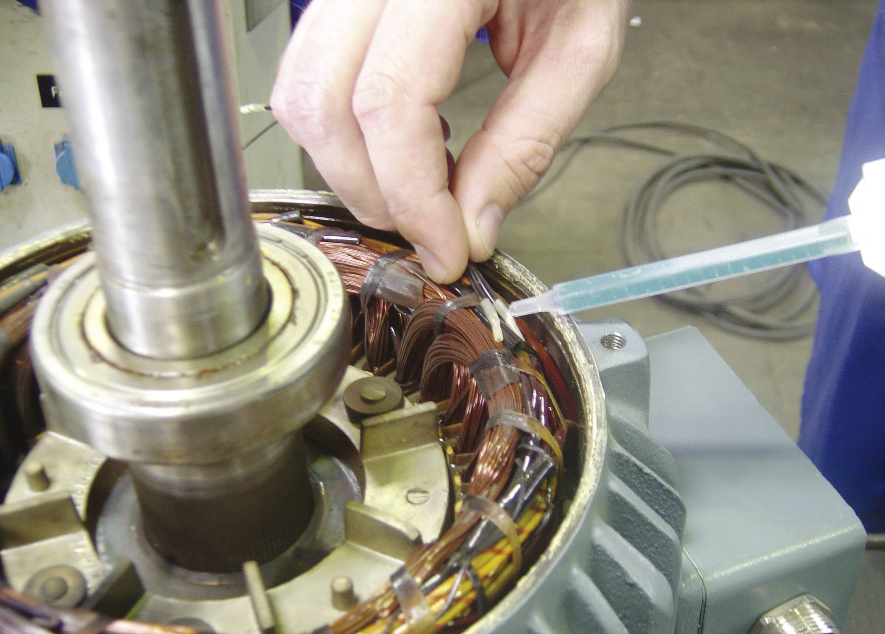 Person repariert einen Elektromotor, injiziert Flüssigkeit in die Kupferspulen. Umgebung deutet auf eine Werkstatt hin.