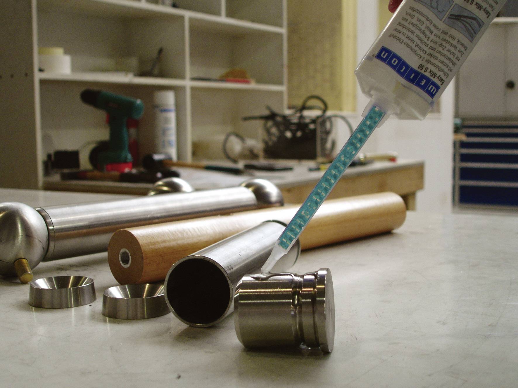 Eine Werkstatt mit verschiedenen Werkzeugen und Materialien. Im Vordergrund sind Metallteile und eine Klebetube zu sehen.