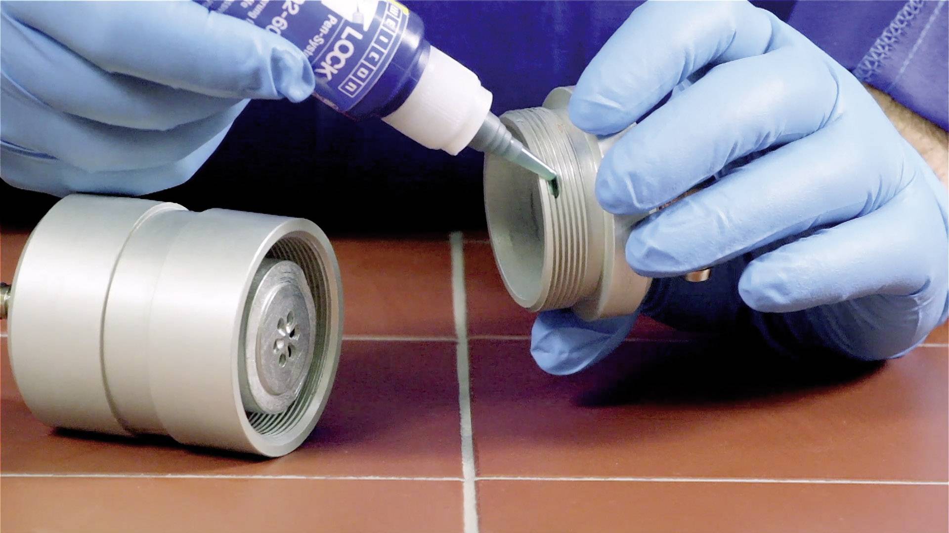 Person in blauen Handschuhen trägt flüssigen Dichtstoff auf Schraubengewinde eines Metallzylinders auf.