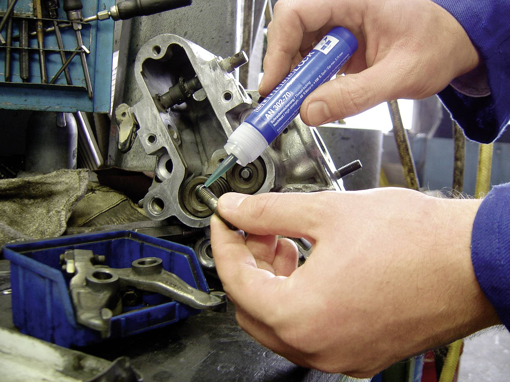 Eine Person trägt mit einer Flasche blauer Flüssigkeit ein Schmiermittel auf ein Metallteil auf, das auf einer Maschinenkomponente befestigt ist.