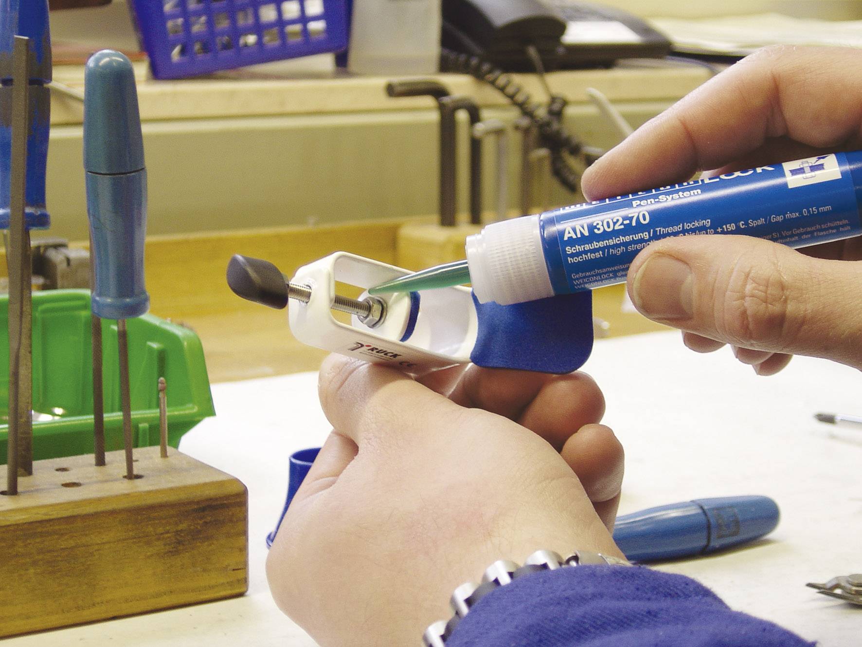 Eine Person trägt blauen Kleber auf einen Gewindebolzen in einer Werkstatt auf, um ihn zu sichern. Im Hintergrund sind Werkzeuge sichtbar.