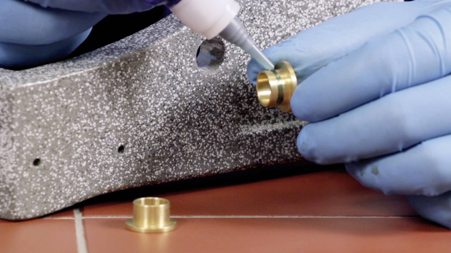 Eine Person mit blauen Handschuhen bringt Kleber auf eine kleine, metallische Komponente an. Im Hintergrund eine strukturierte Oberfläche.