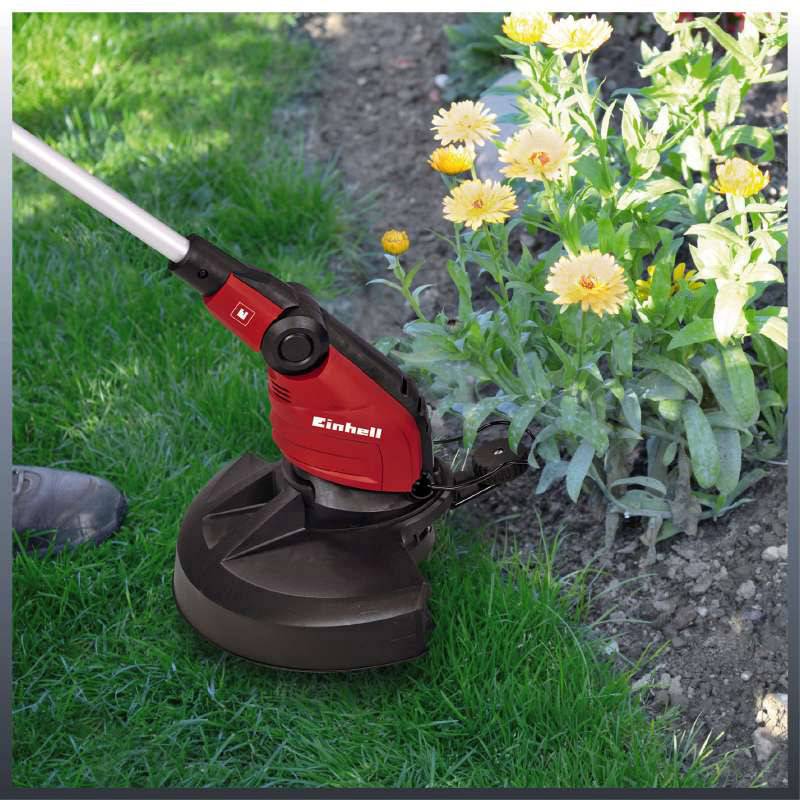 Eine Person hält einen roten Rasentrimmer, trimmt den Rand eines Rasens neben blühenden gelben Blumen im Garten.
