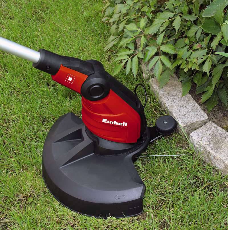 Roter Rasentrimmer schneidet Gras entlang eines Gartenrands neben grünen Pflanzen und Steinen.