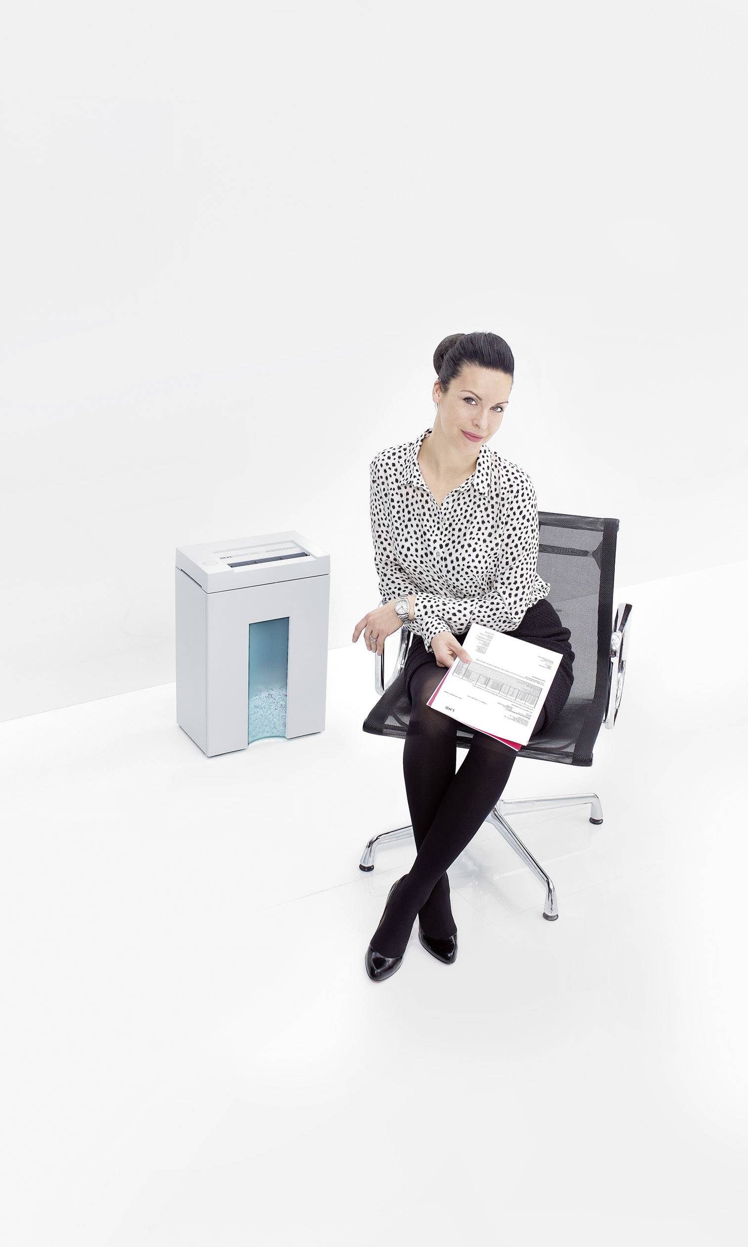 Eine Frau sitzt auf einem Bürostuhl neben einem Aktenvernichter und hält Papiere in der Hand. Der Hintergrund ist schlicht weiß.