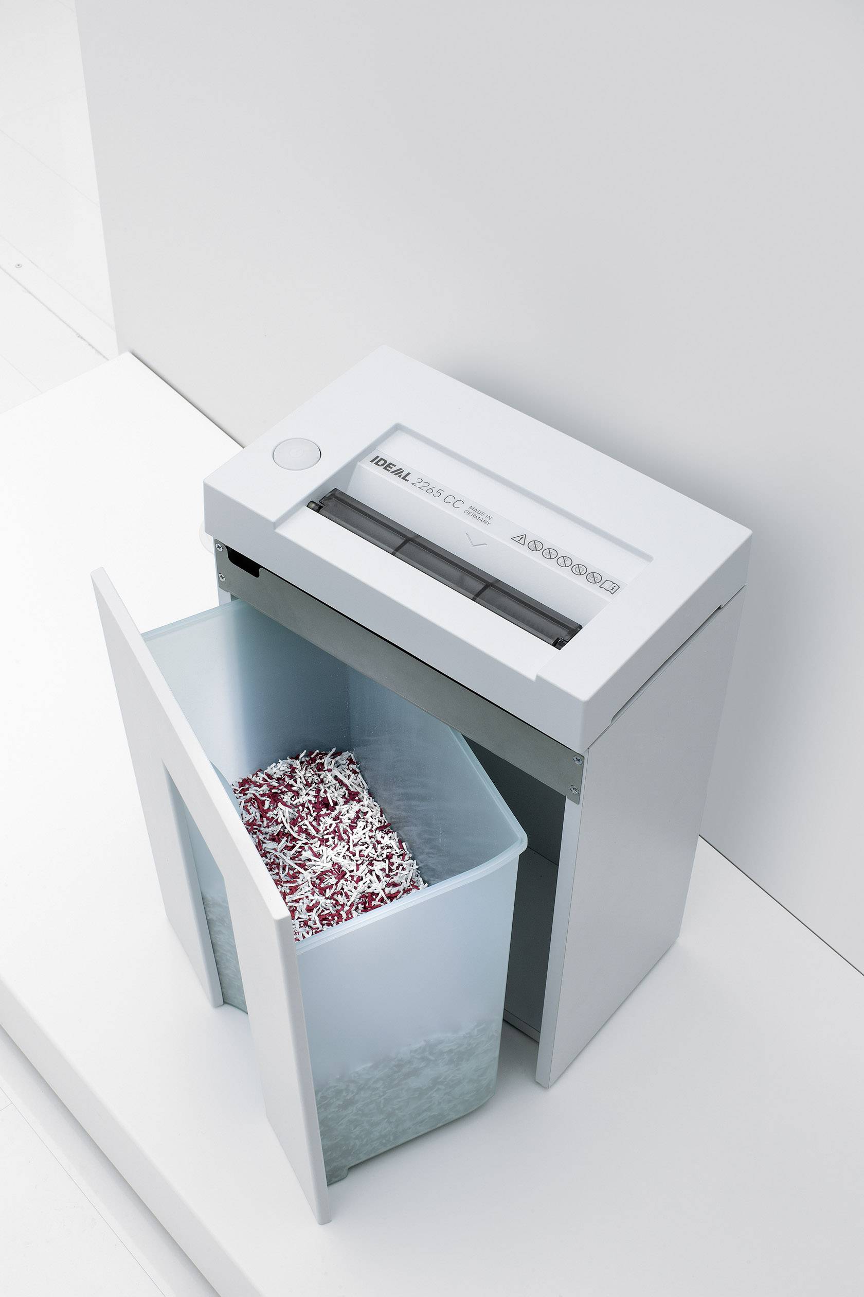 Ein Aktenvernichter mit offenem Papierauffangbehälter, gefüllt mit zerkleinerten Papierstücken. Gerät steht auf einem weißen Boden.