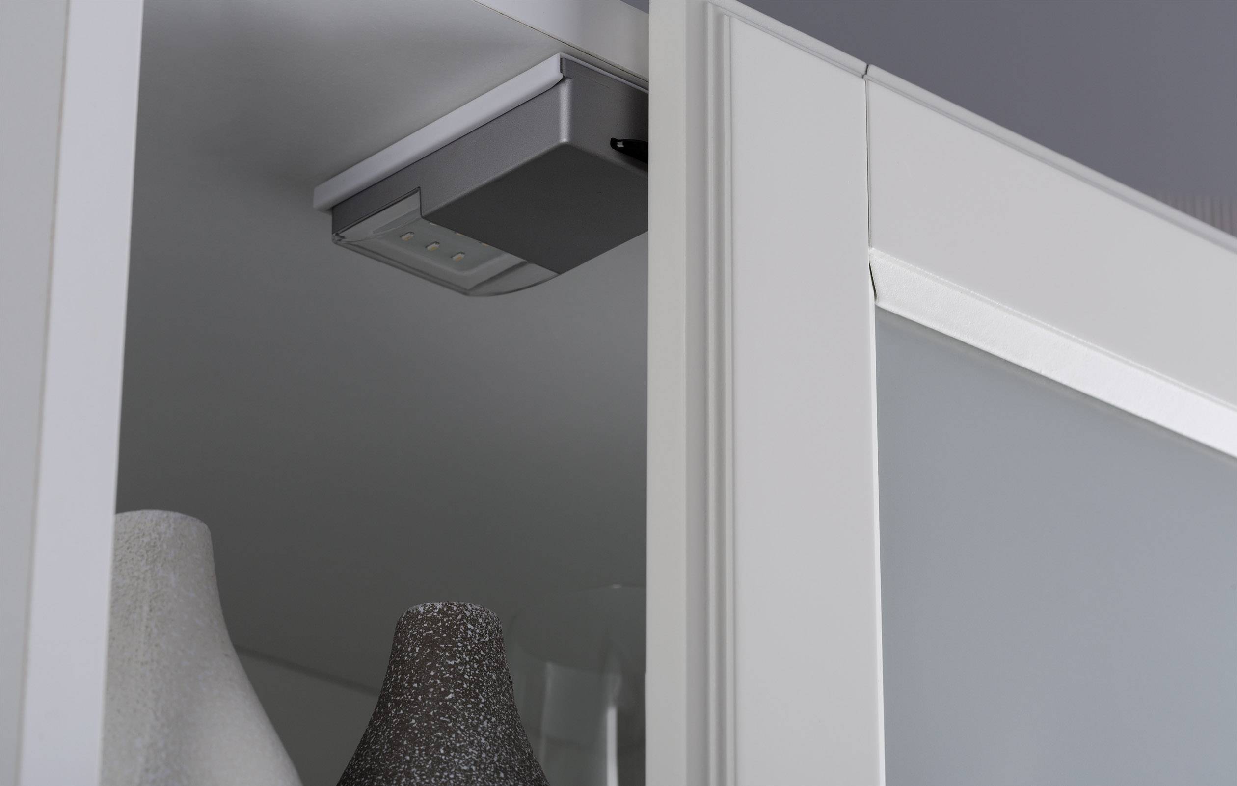 Ein Schrank mit einer eingebauten LED-Leuchte an der Innenseite der oberen Kante und Vasen im Hintergrund.