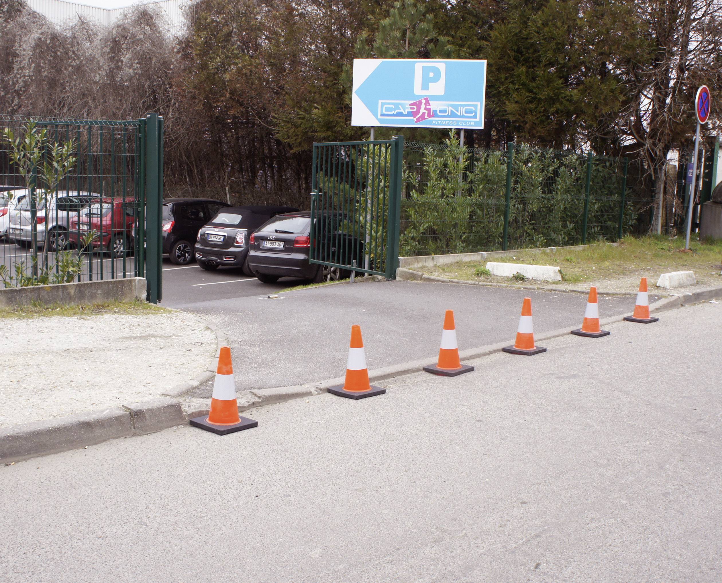 Autos fahren durch ein geöffnetes Tor in einen Parkplatz ein. Ein blaues Schild mit 'Parking' weist den Weg. Verkehrskegel stehen am Rand.