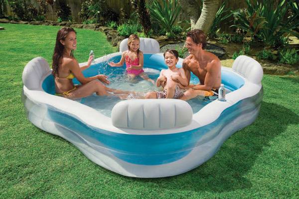 Eine Familie mit zwei Kindern sitzt in einem aufblasbaren Pool im Garten. Alle sind lachend und scheinen die sonnige Atmosphäre zu genießen.