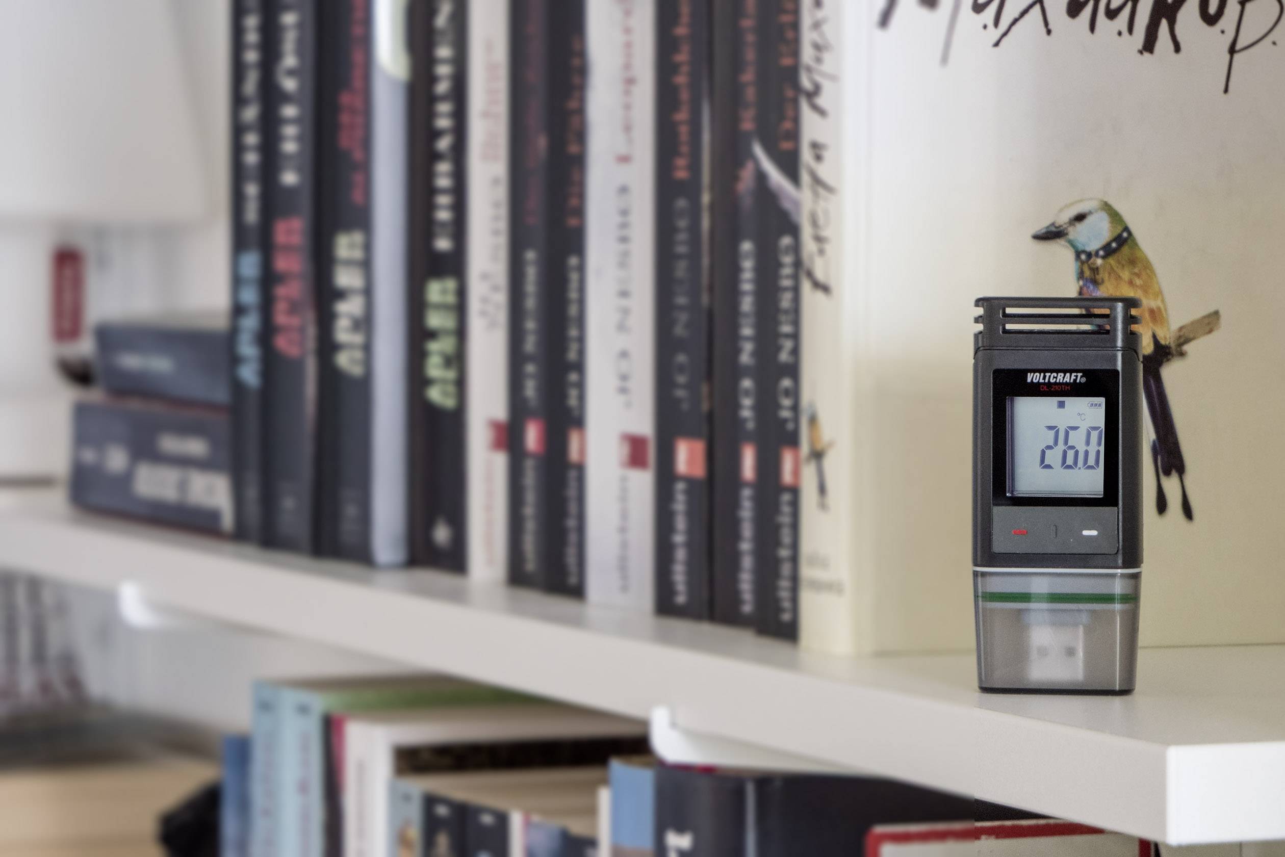 Ein digitales Hygrometer zeigt '26.0' auf einem Bücherregal an, umgeben von verschiedenen Büchern.