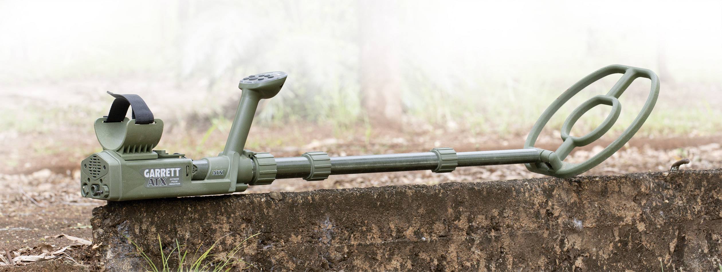 Ein grüner Metalldetektor liegt auf einer Betonoberfläche im Freien. Der Hintergrund zeigt unscharfe Bäume und Gras.