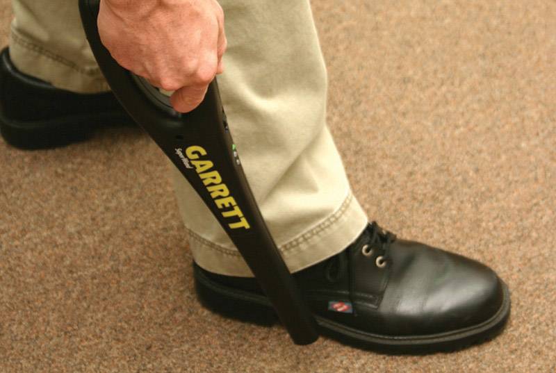 Eine Person scannt mit einem Hand-Metalldetektor, der in der rechten Hand gehalten wird, den Bereich um ihren linken Schuh.