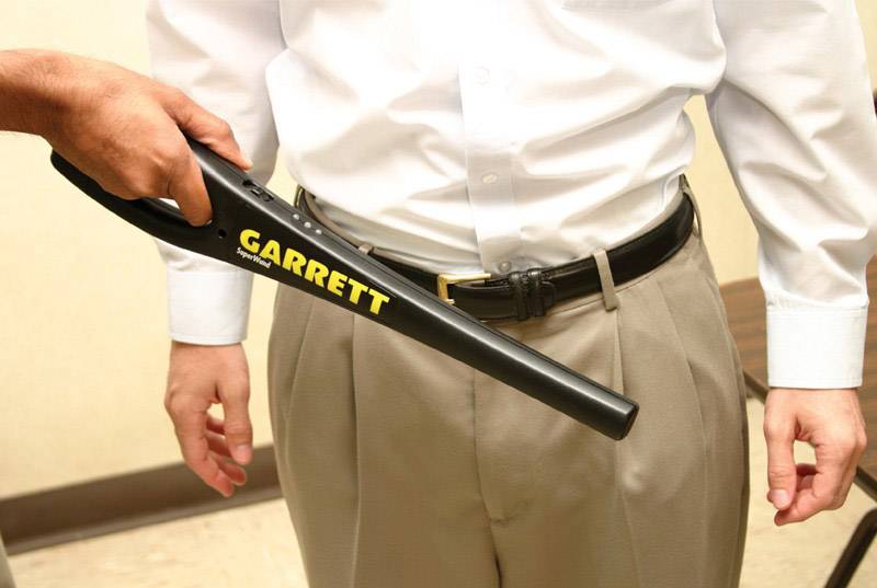 Eine Person wird mit einem Hand-Metalldetektor auf Metallgegenstände überprüft. Der Detektor ist schwarz und von der Marke Garrett.
