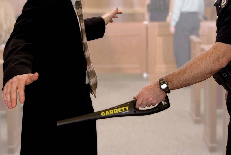 Eine Sicherheitskontrolle in einem Gerichtssaal. Ein Beamter scannt mit einem Metalldetektor eine Person.