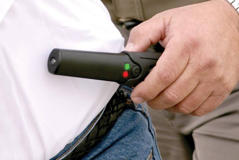 Eine Person hält ein handheld Metalldetektor über der Hosentasche einer anderen Person. Der Detektor zeigt grüne und rote Lichter.