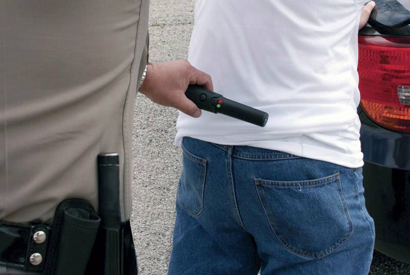 Ein Polizeibeamter durchsucht eine stehende Person mit einem Handmetalldetektor. Die Person ist in Freizeitkleidung neben einem Auto.