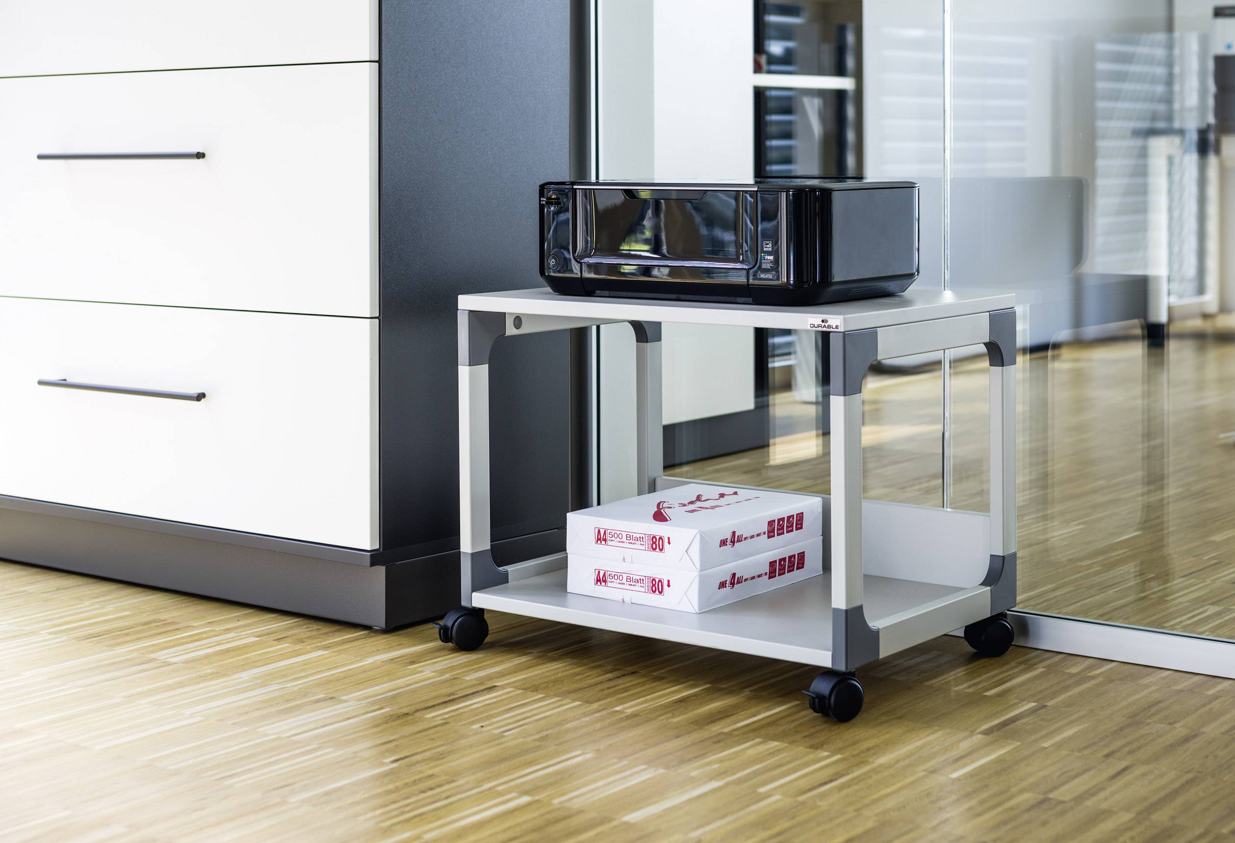 Ein Drucker steht auf einem rollbaren Ablagetisch in einem modernen Büro, darunter liegen zwei Stapel Druckerpapier.