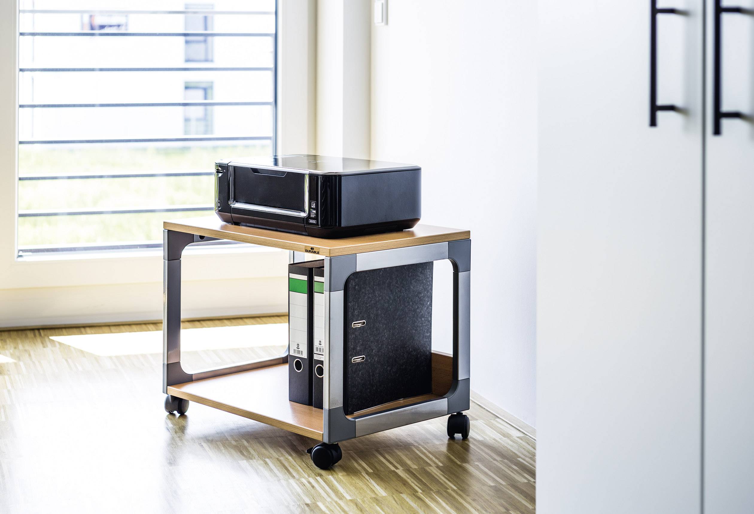 Drucker auf einem kleinen, beweglichen Tisch mit drei Ordnern darunter, in einem hellen, modernen Büro mit Holzfußboden.