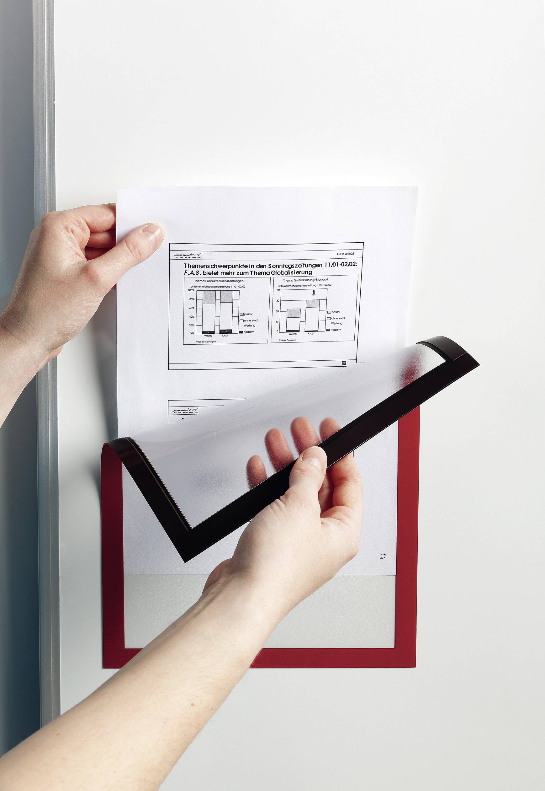 Zwei Hände halten ein Blatt Papier mit einem Diagramm, das in einem transparenten Folienschutz mit rotem Rahmen an einer Wand angebracht wird.