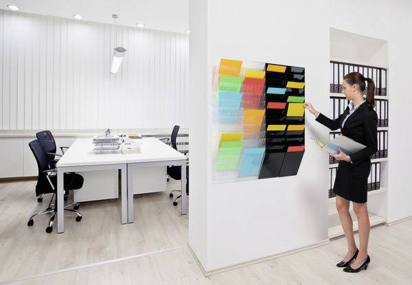 Eine Frau in Geschäftskleidung steht vor Wandregalen in einem modernen Büro, hält Ordner und entnimmt ein Dokument.