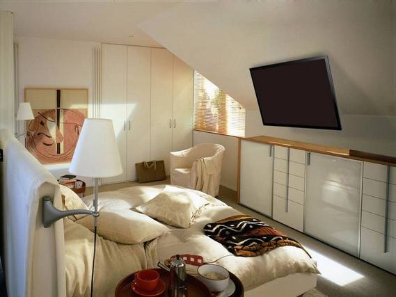 Ein gemütliches Schlafzimmer mit einem großen Doppelbett, einem Sessel, einem Flachbildfernseher an der Wand und einem Fenster mit Lamellenjalousien.