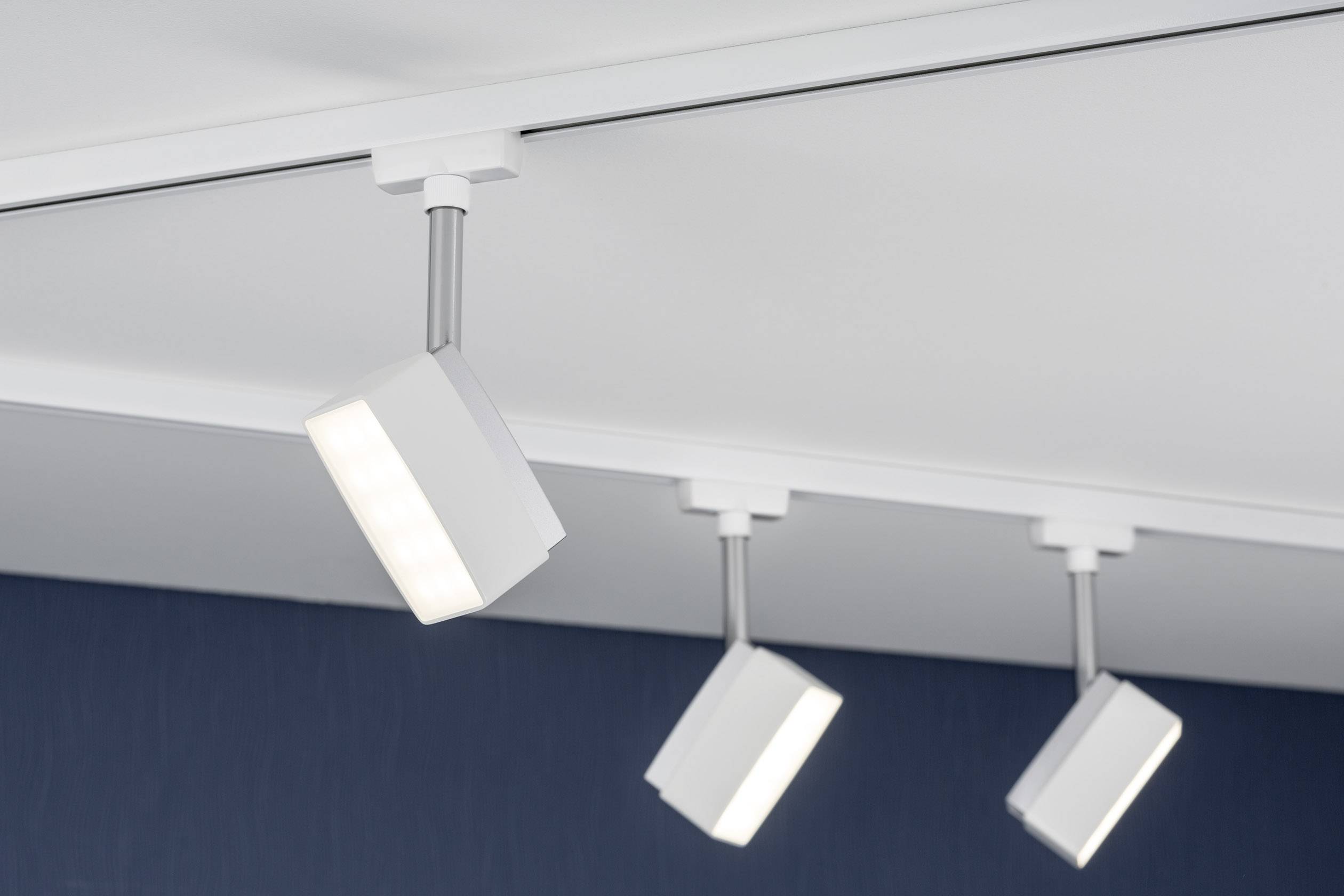 Drei weiße, quadratische Deckenstrahler an einer Schiene montiert, leuchten nach unten. Der Hintergrund ist dunkelblau.