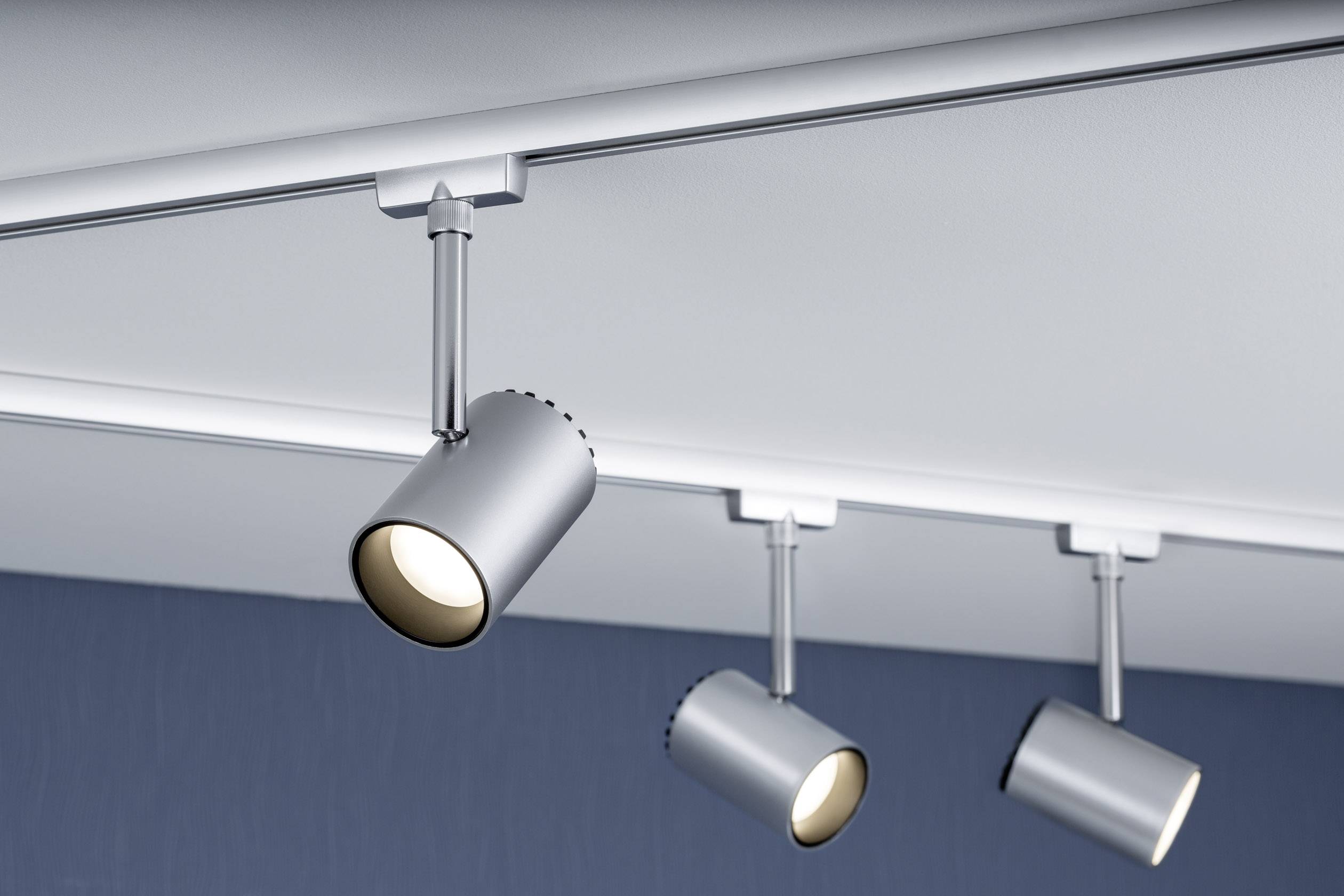 Deckenleuchte mit drei silbernen, zylinderförmigen Spots, die auf einer Schiene montiert sind und auf einen blauen Hintergrund leuchten.