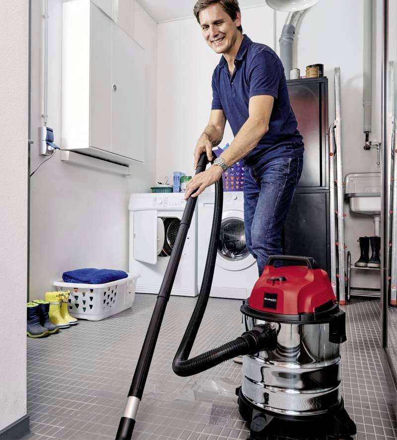 Ein Mann in einem Hauswirtschaftsraum saugt mit einem Nass-Trockensauger den Boden. Im Hintergrund sind eine Waschmaschine und ein Korb mit Wäsche zu sehen.