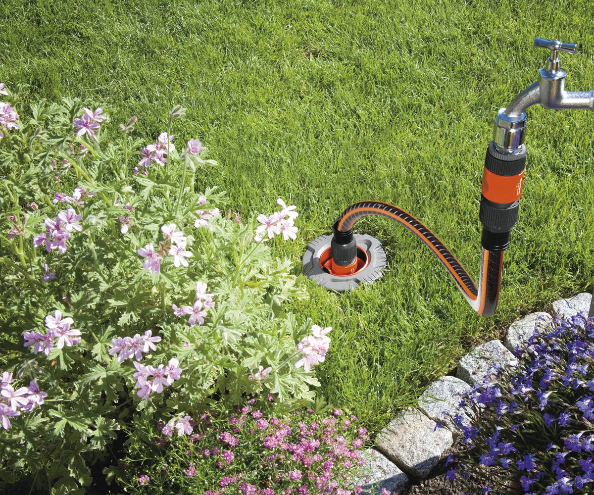 Ein Gartenschlauch mit Aufsatz bewässert den Rasen neben einem Blumenbeet mit rosa und lila Blüten, umgeben von grünen Pflanzen.