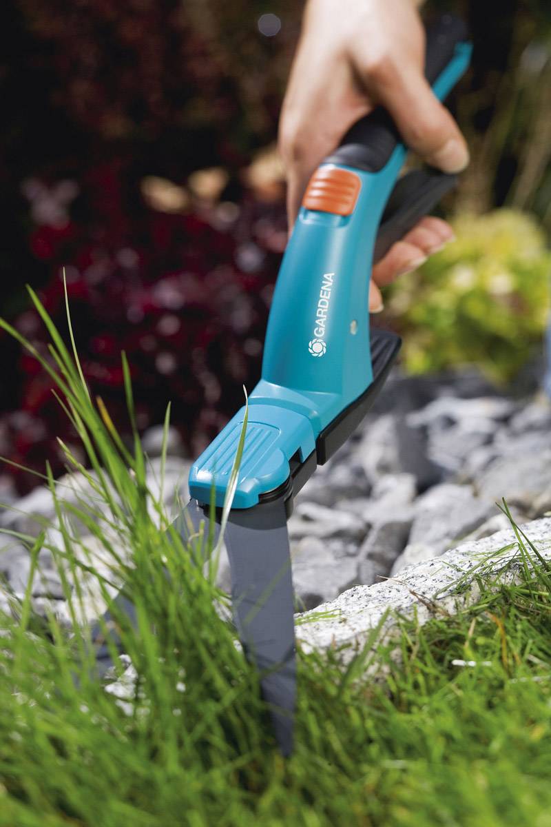 Eine Hand schneidet mit einer blauen und schwarzen Grasschere Gras entlang eines steinigen Pfades im Garten.