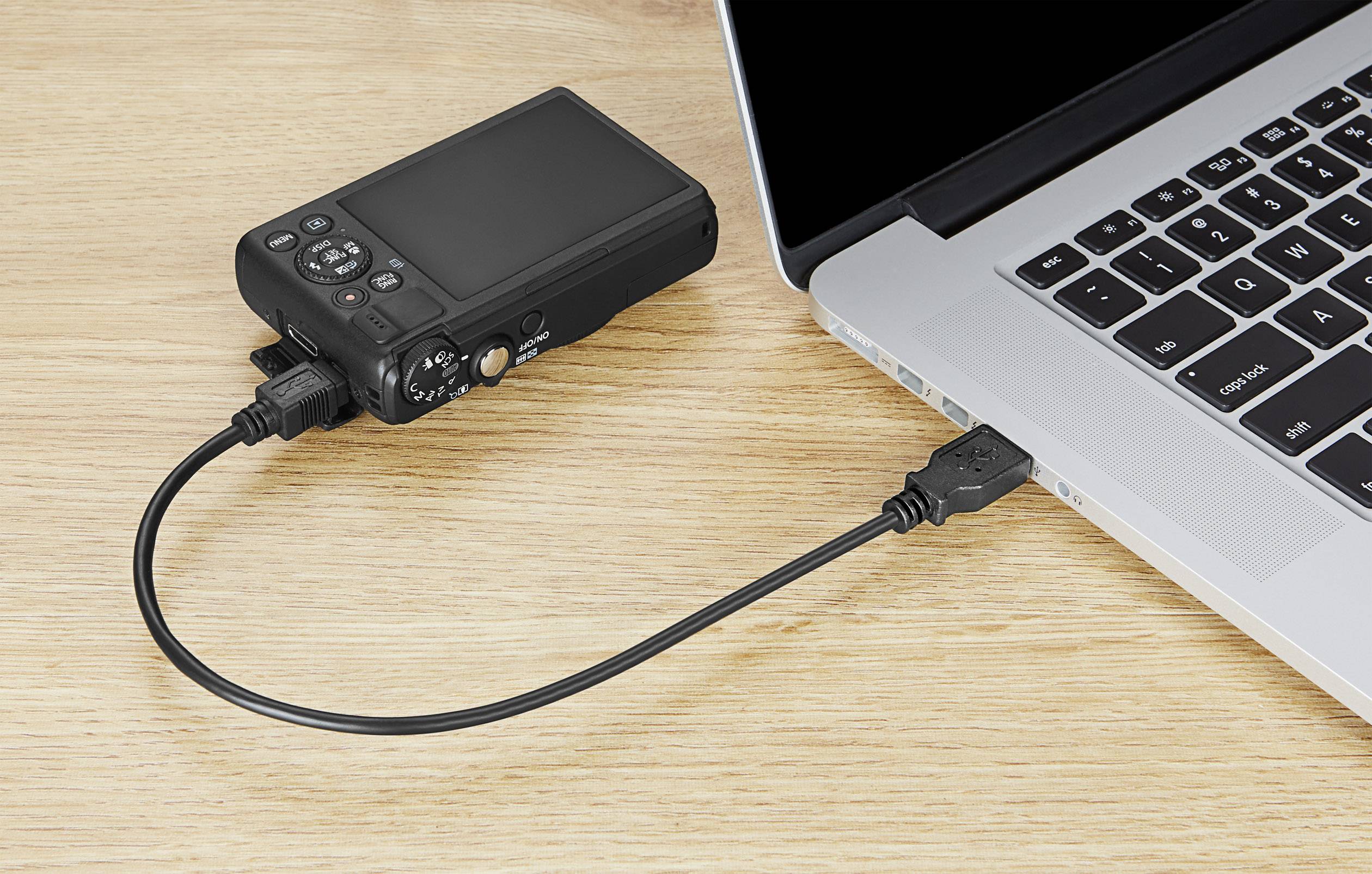 Eine Digitalkamera ist über ein USB-Kabel mit einem Laptop verbunden. Der Laptop zeigt eine teilweise geöffnete Tastatur.