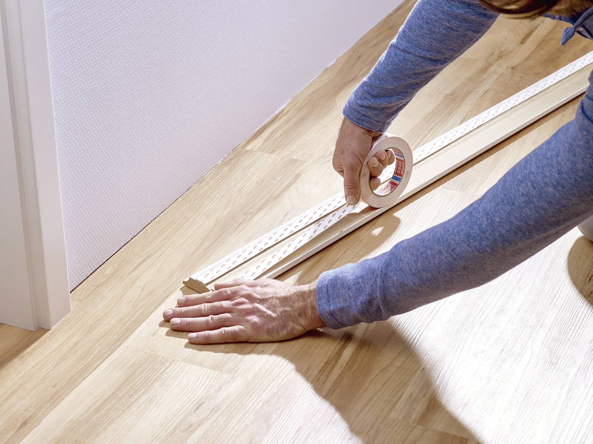 Person befestigt mit Klebeband eine Holzleiste an einer hellen Wand auf einem Holzboden.