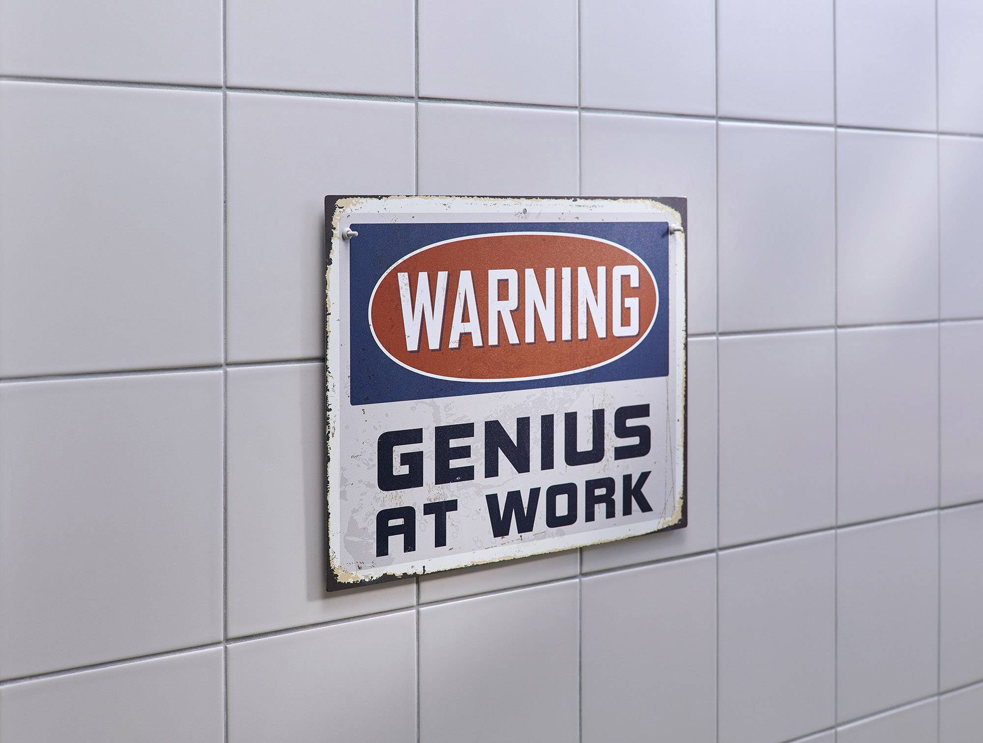 Warnschild an einer gefliesten Wand mit der Aufschrift 'Warning: Genius at Work'.