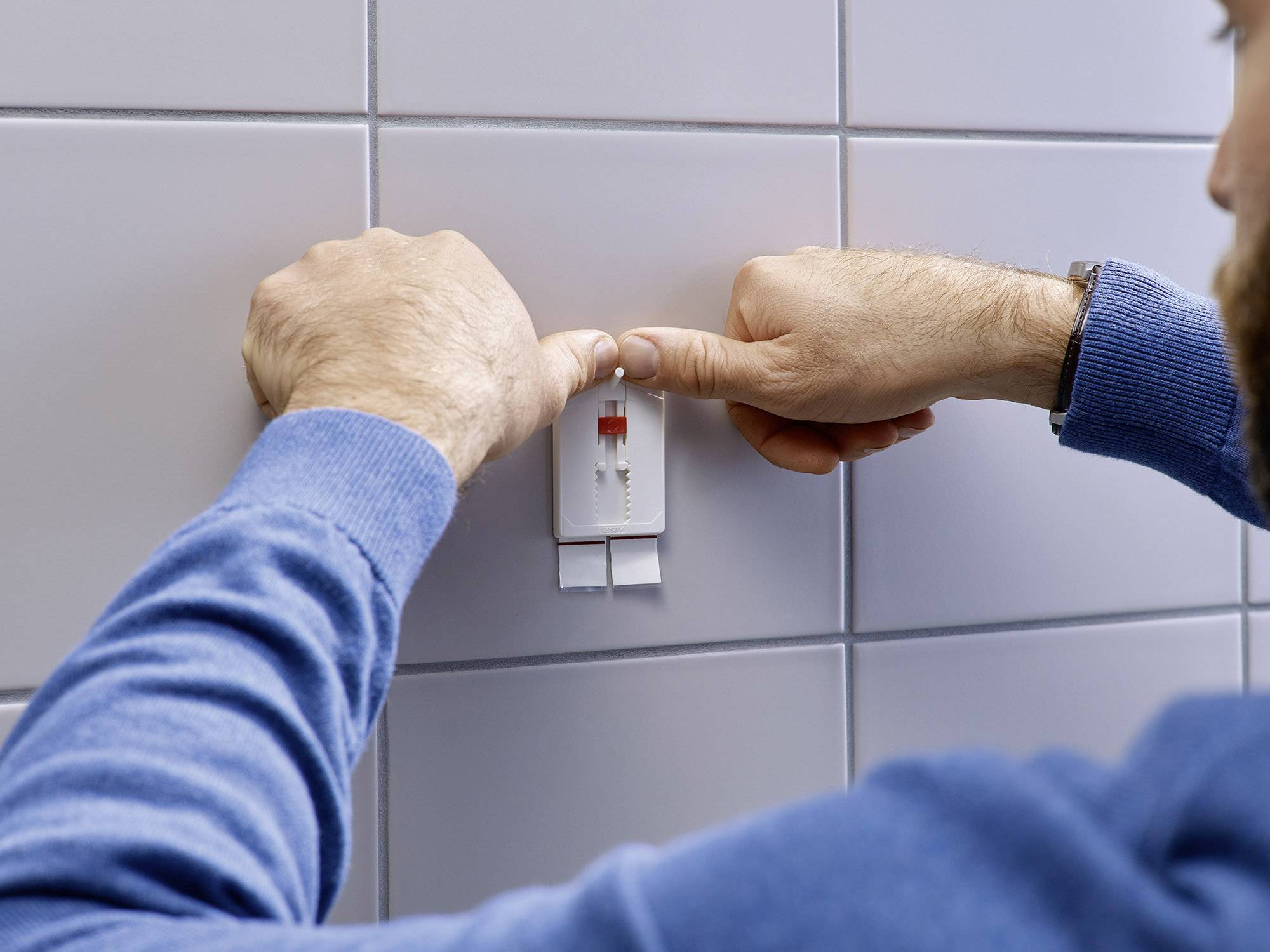 Eine Person bezieht doppelseitiges Klebeband von einer weißen Rolle, die an einer gefliesten Wand angebracht ist.