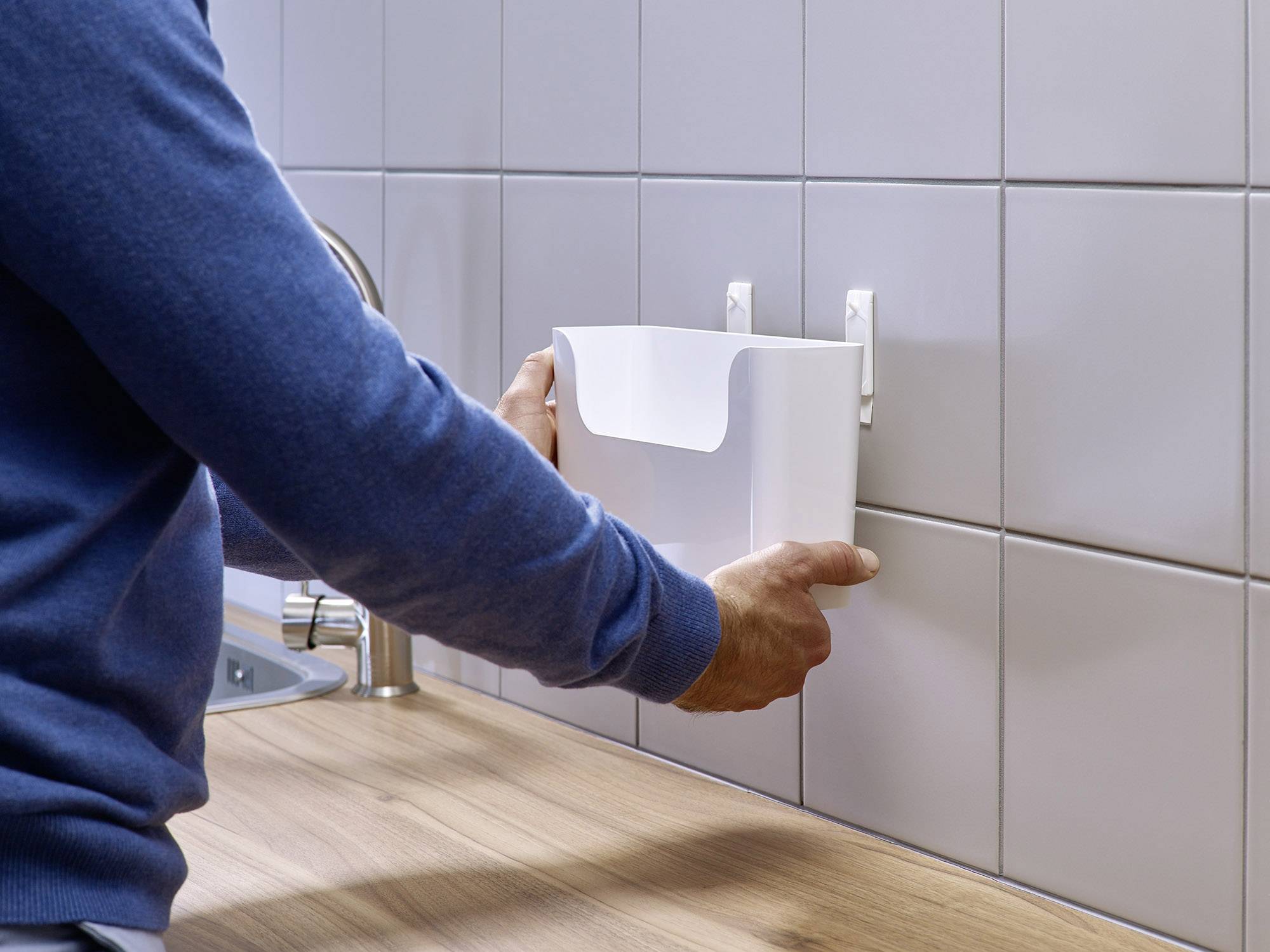 Eine Person montiert einen weißen, hängenden Papierkorb an einer gefliesten Wand in der Nähe eines Spülbeckens auf einer Holzarbeitsplatte.