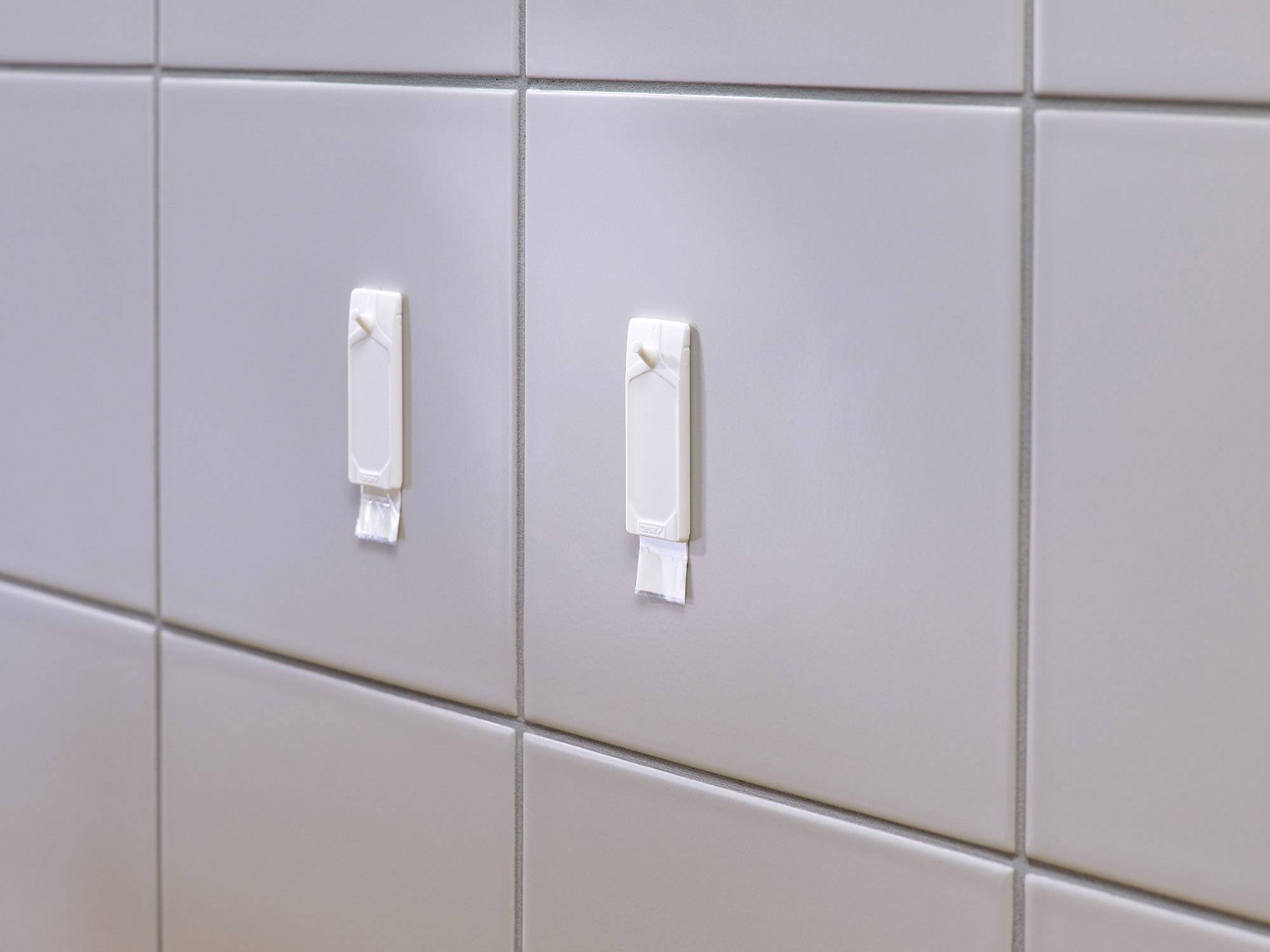 Zwei weiße Haken auf hellgrauen Fliesen, befestigt mit Klebestreifen. Ideal zur Anbringung von leichten Gegenständen im Badezimmer.