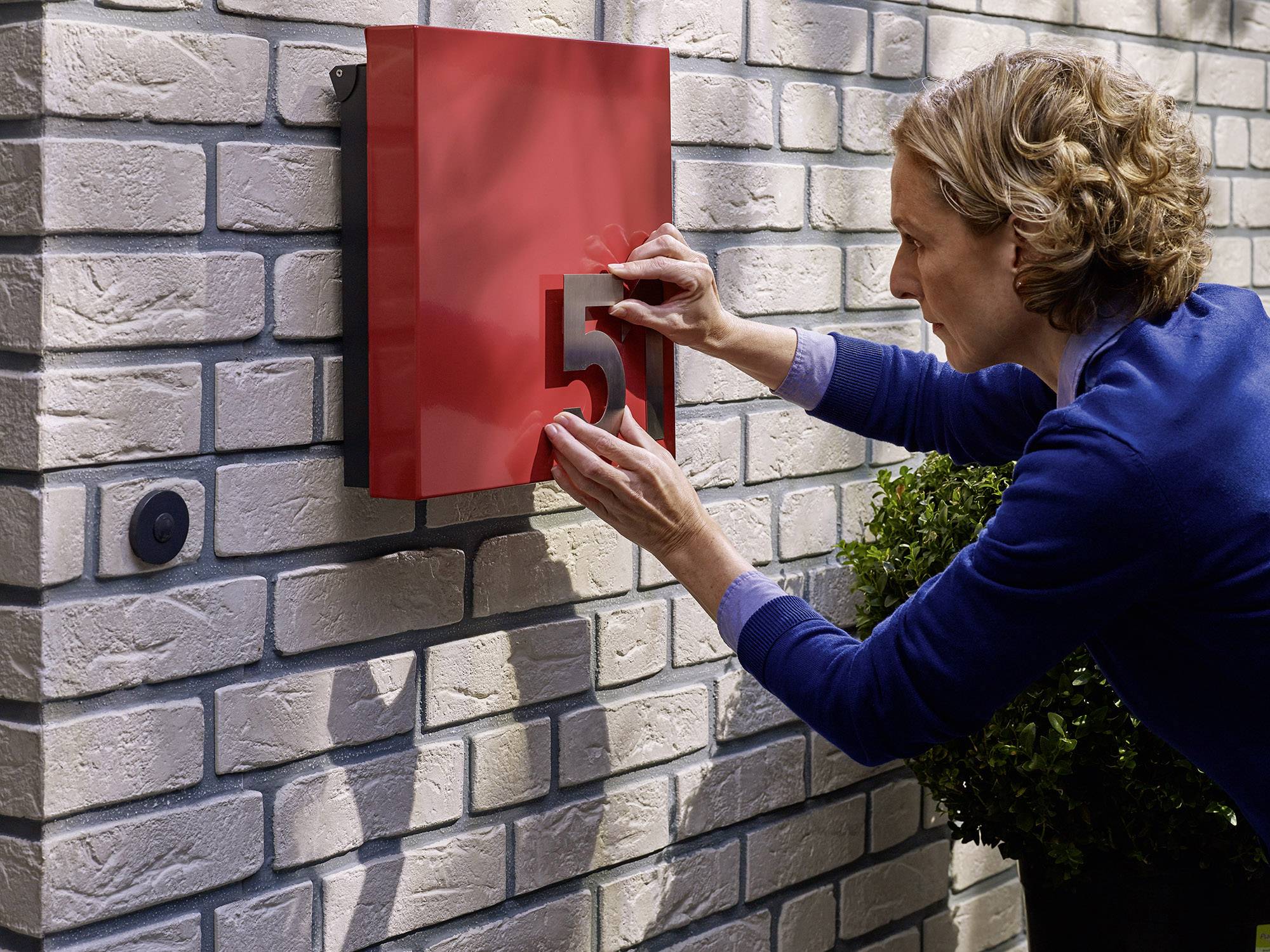 Eine Frau befestigt die Zahl '5' an einem roten Schild an einer weißen Ziegelwand neben einer Eingangstür.