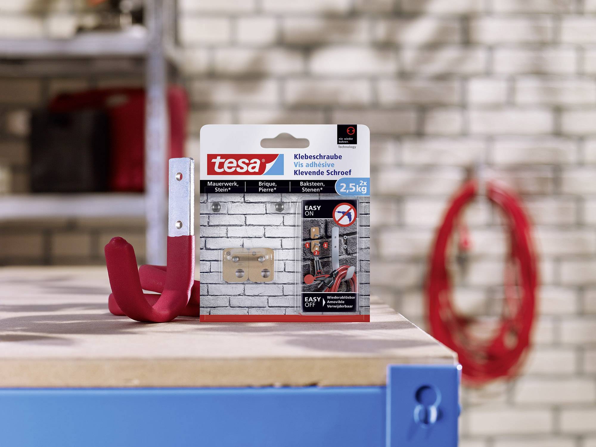 Packung mit tesa-Klebeschrauben auf einem Tisch vor Ziegelmauer. Die Schrauben sind für feste Oberflächen geeignet und können bis zu 2,5 kg tragen.