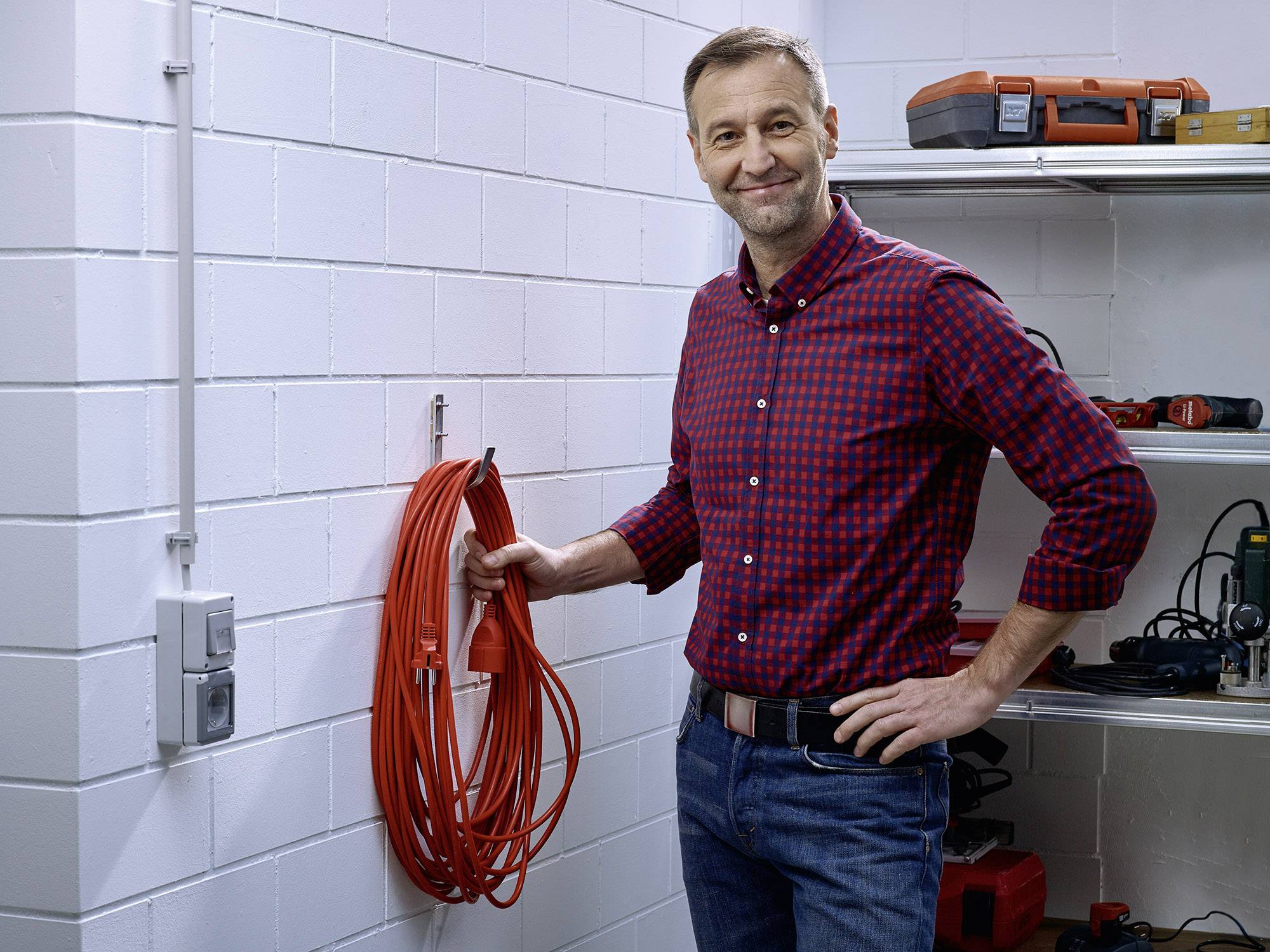 Ein lächelnder Mann steht in einer Werkstatt neben einem aufgerollten roten Kabel an der Wand. Im Hintergrund sind Werkzeuge auf Regalen.