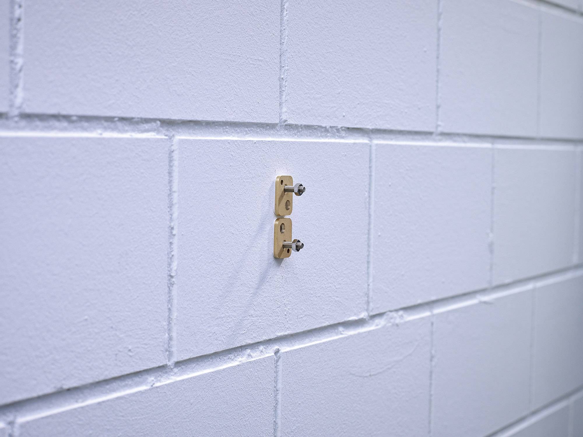 Weiße Ziegelwand mit zwei hervorstehenden Schrauben in einer kleinen Halterung, möglicherweise zur Befestigung eines Gegenstandes.