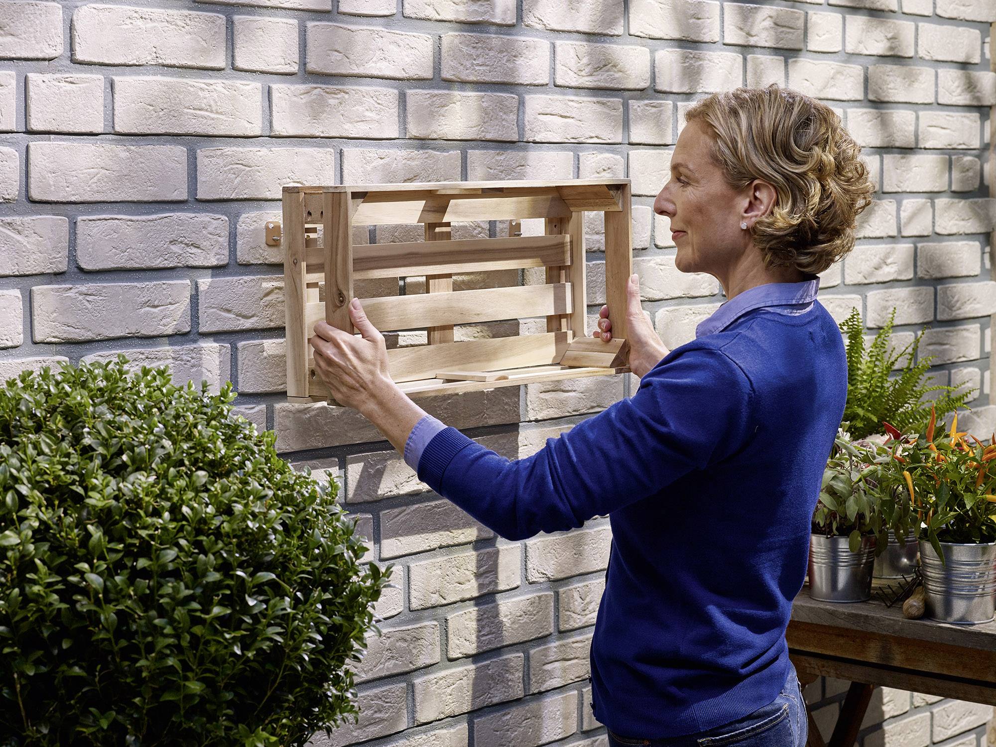 Person montiert hölzernes Regal an eine Ziegelwand im Garten. Pflanzen in der Nähe vermitteln eine natürliche Atmosphäre.