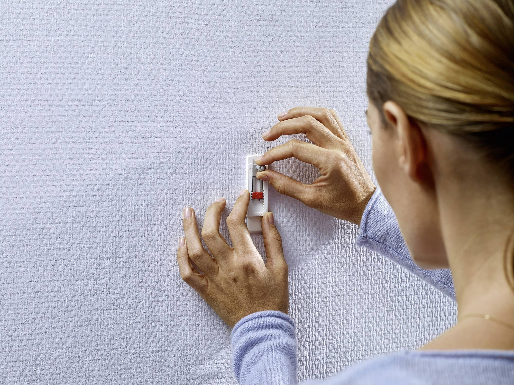 Eine Person befestigt einen kleinen Gegenstand an einer strukturierten Wand, indem sie ihn drückt. Die Hände sind im Fokus.