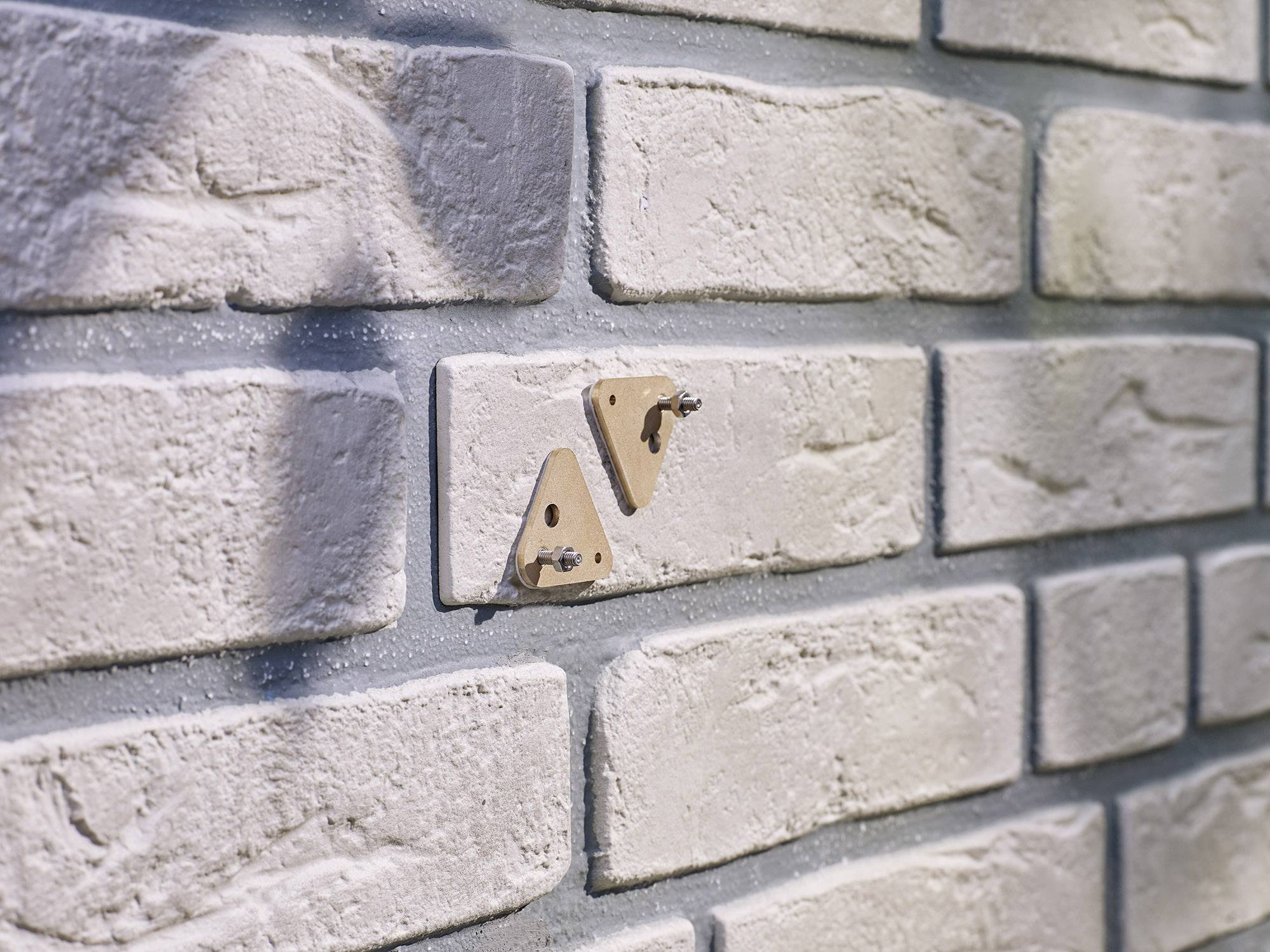 Zwei kleine Holz-Dreiecke, die an einer beige gestrichenen Ziegelwand befestigt sind, werfen Schatten auf die Mauer.
