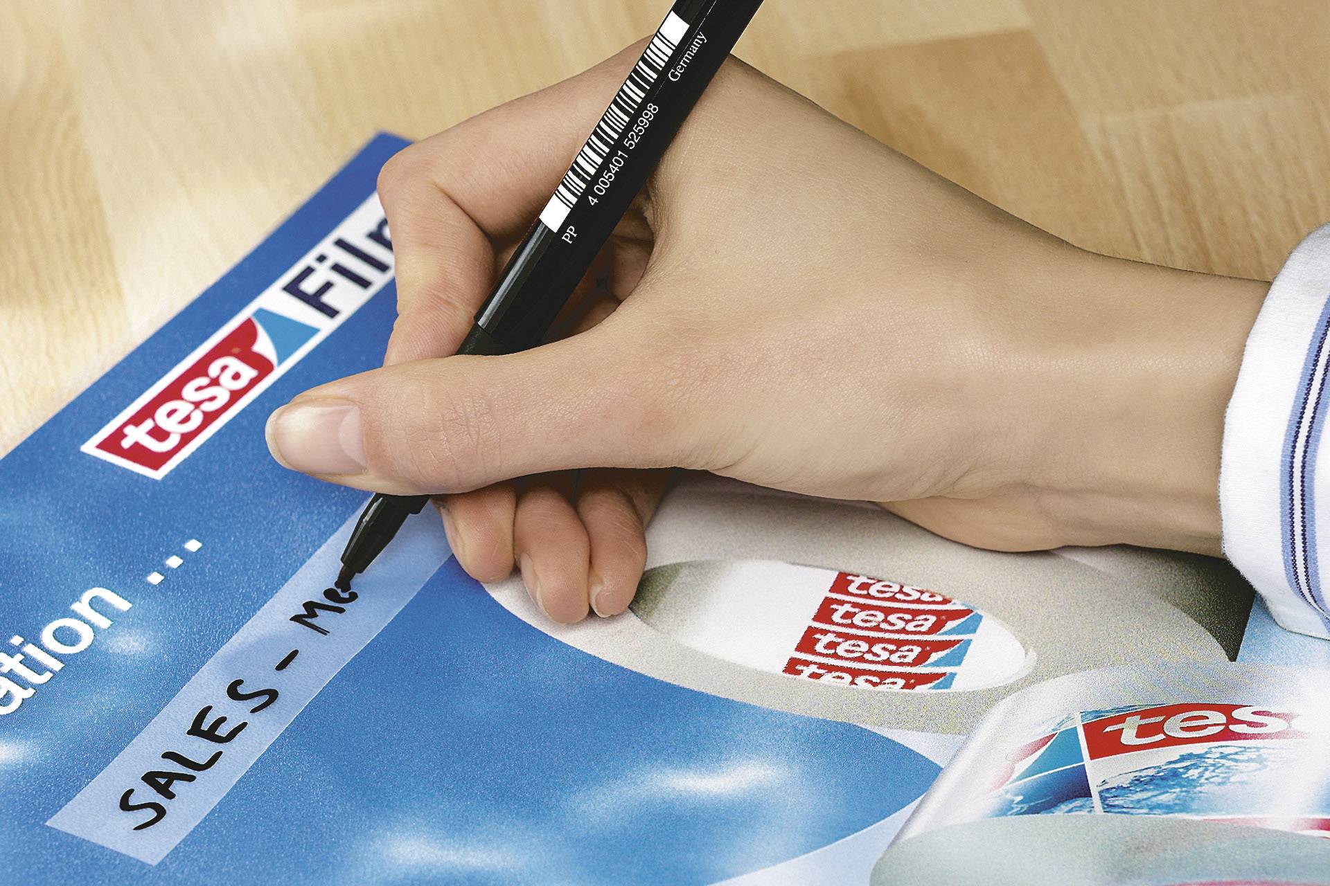 Eine Hand schreibt mit einem schwarzen Stift 'SALES-M' auf blaues Papier mit tesa-Logo, neben einer Rolle Tesa-Klebeband.