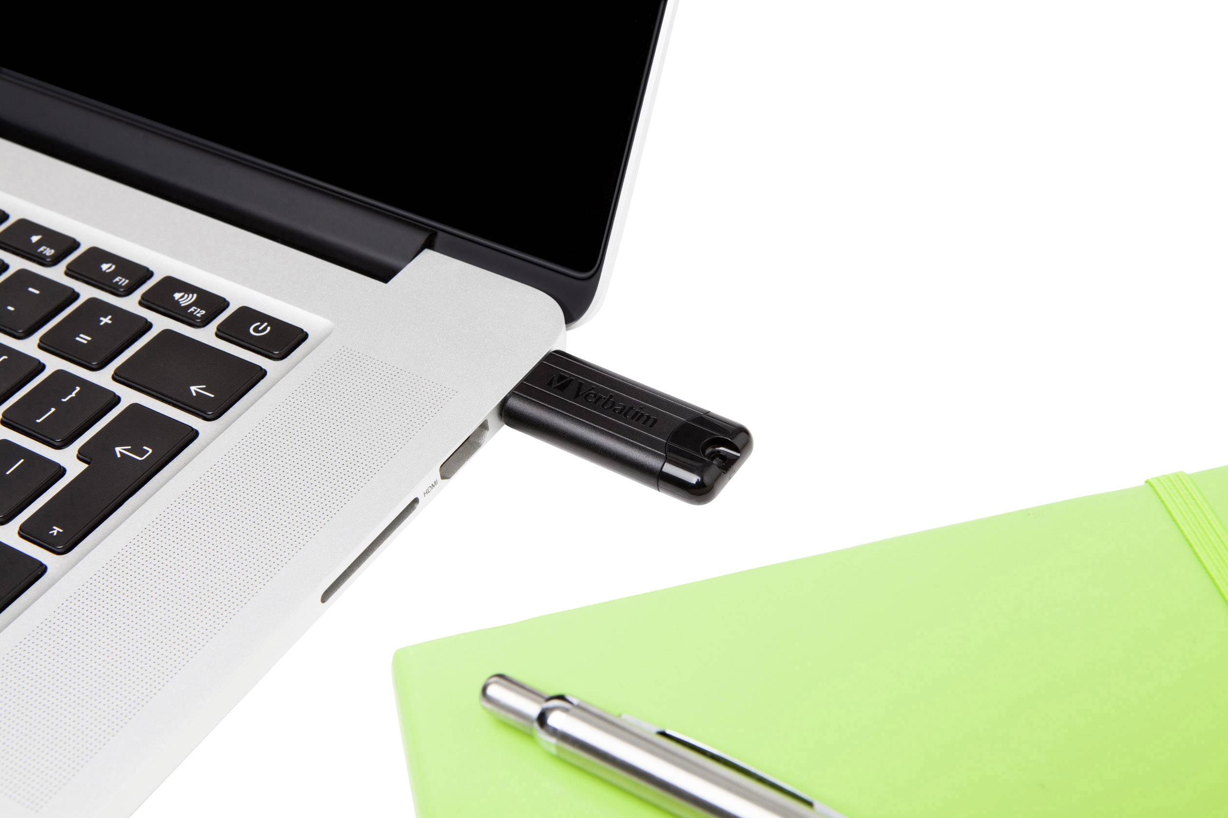 Ein Laptop mit angeschlossenem USB-Stick neben einem grünen Notizbuch und einem silbernen Kugelschreiber auf einem weißen Hintergrund.